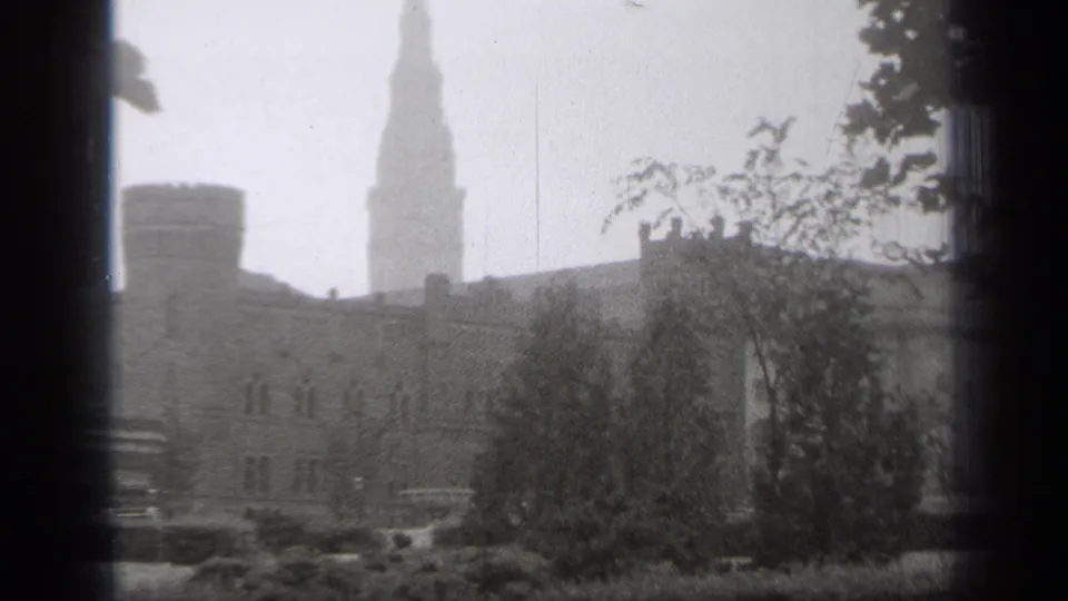 Soft black-and-white Cleveland frame with a steeple, older building facade, trees, shrubs, and parked period vehicles.