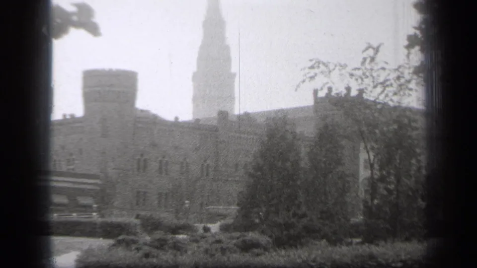1938 Cleveland exterior with a hazy steeple above masonry buildings, leafy trees, shrubs, and a parked vehicle.