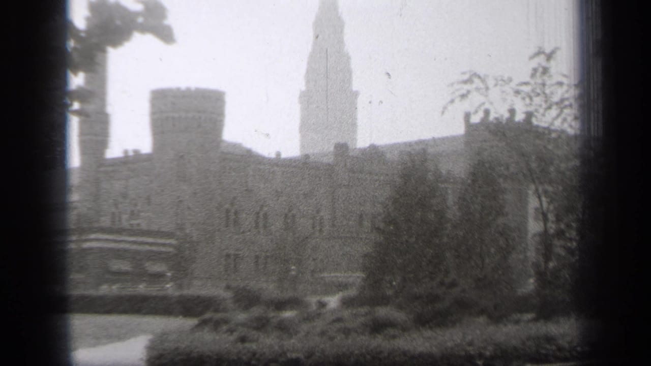 Stone building with turret and tall spire seen through trees in black-and-white film.