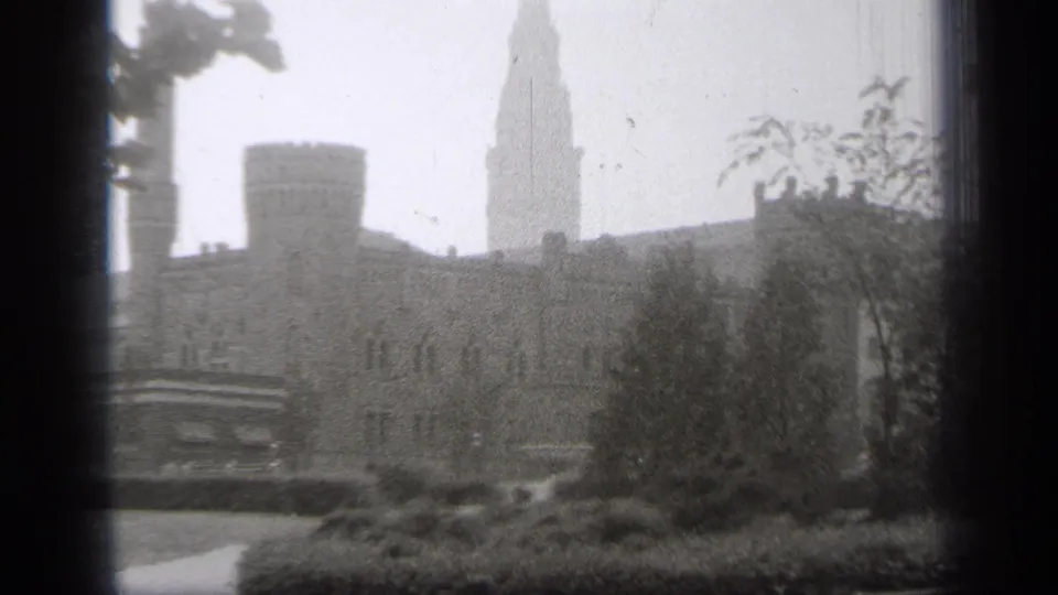 1938 Cleveland 8mm frame showing a stone building facade, crenellated turret, and tall spire partly obscured by trees.