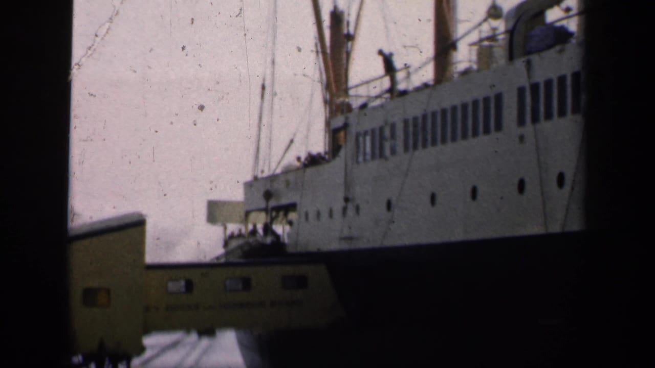 1949 England home-movie frame showing a large ocean liner at port with a yellow gangway and distant dockside figures.