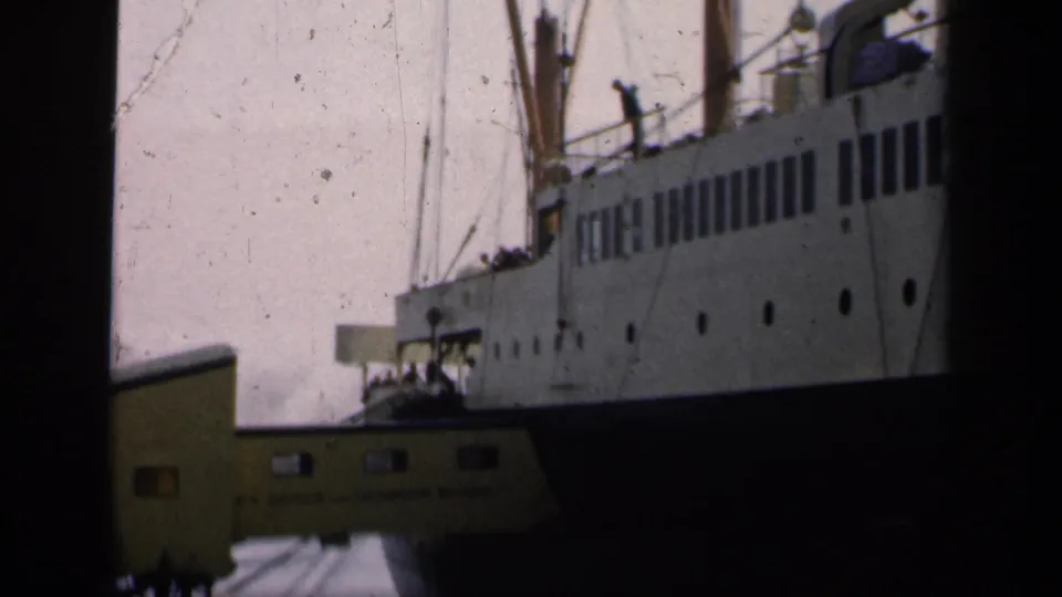 1949 England home-movie frame showing a large ocean liner at port with a yellow gangway and distant dockside figures.