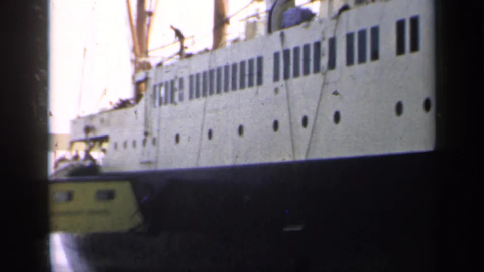 1949 England home-movie frame of a large passenger ship side, showing rows of windows, portholes, rigging, and the gangway below.
