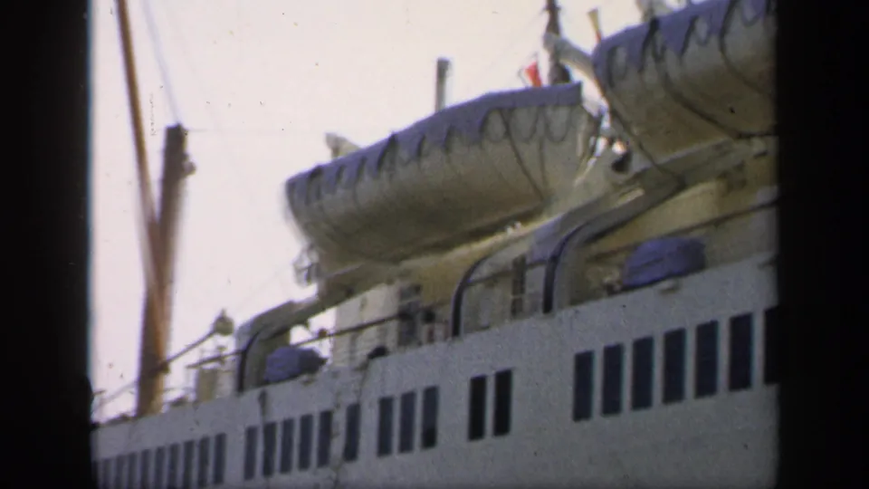 1949 England home-movie frame of a passenger ship section with covered lifeboats, railing, rigging, and rows of dark windows.
