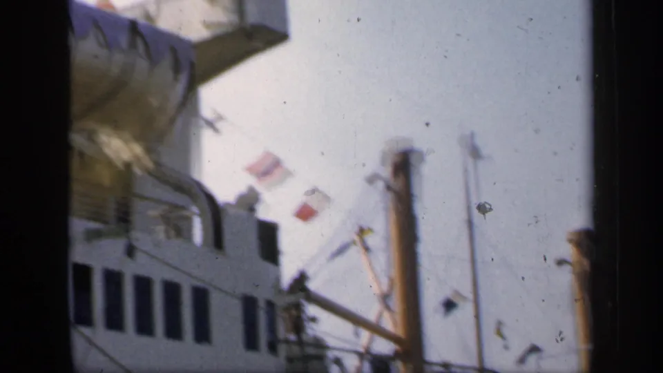 1949 England 8mm frame showing ship windows, railings, smokestacks, mast lines, and small flags including a French flag against a bright sky.