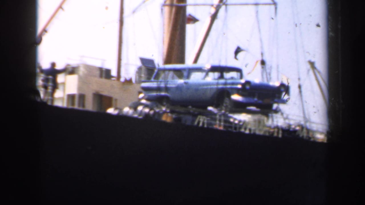 A blue mid-century station wagon appears suspended above or near a cargo ship deck in 1949 England footage.