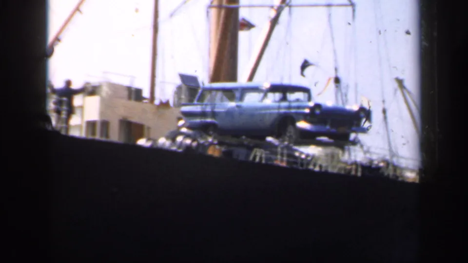 A blue mid-century station wagon appears suspended above or near a cargo ship deck in 1949 England footage.