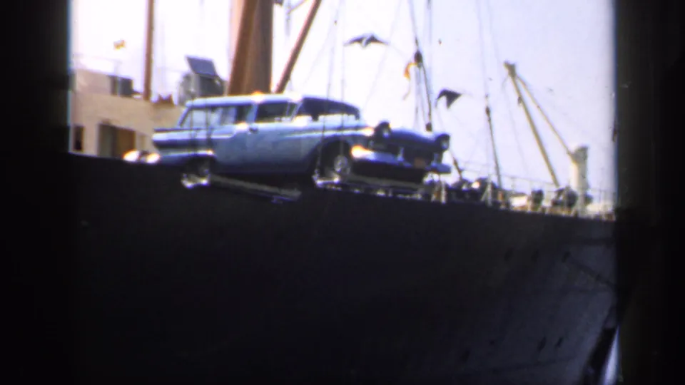 A blue station wagon is visible on a cargo ship deck in 1949 England, surrounded by rigging, flags and ship structures.