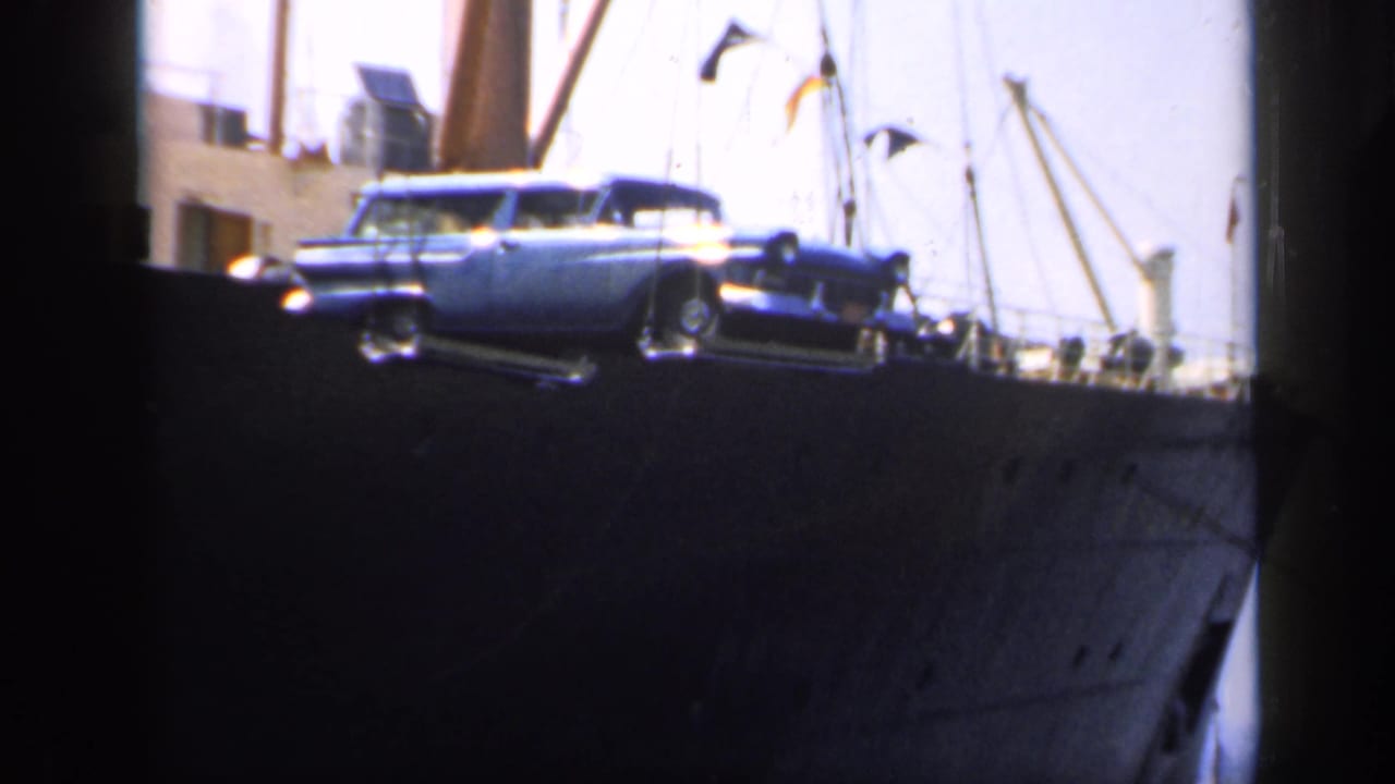 Blue estate car suspended beside a dark ship hull at a harbor.