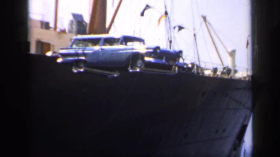 In 1949 England footage, a blue estate car hangs from crane gear beside a large ship with rigging and deck figures visible.