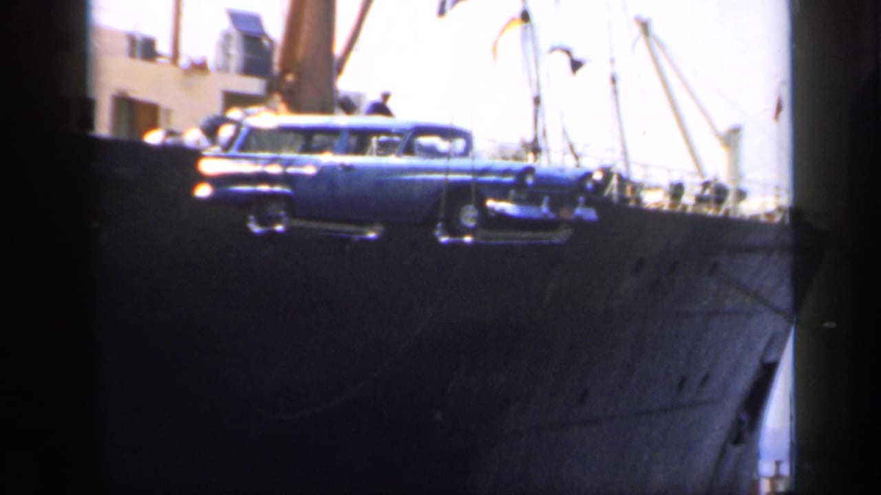 Suspended blue car hangs near the rail of a large ship.