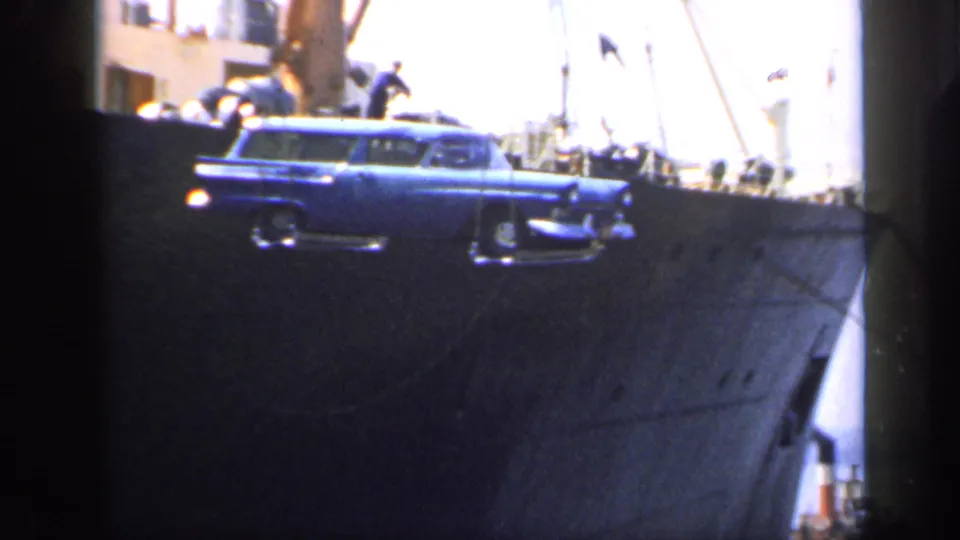 A blue station wagon is seen closer to the ship's side, suspended by rigging during a dockside loading operation in England.