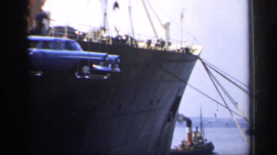 The suspended car moves partly out of frame near the ship bow, with harbor water, rigging lines, and a small workboat visible below.