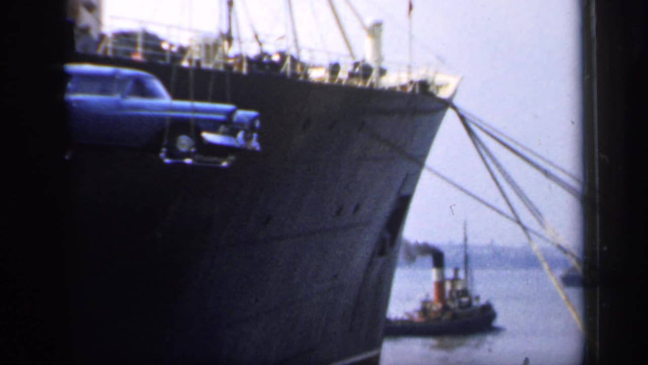 Blue station wagon suspended by crane beside a large dark ship hull with a tugboat in the background.