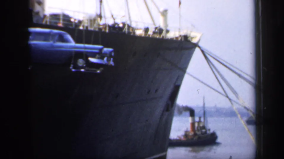 In England in 1949, a blue station wagon hangs in a crane sling beside a large ship while a small tugboat moves near the harbor.