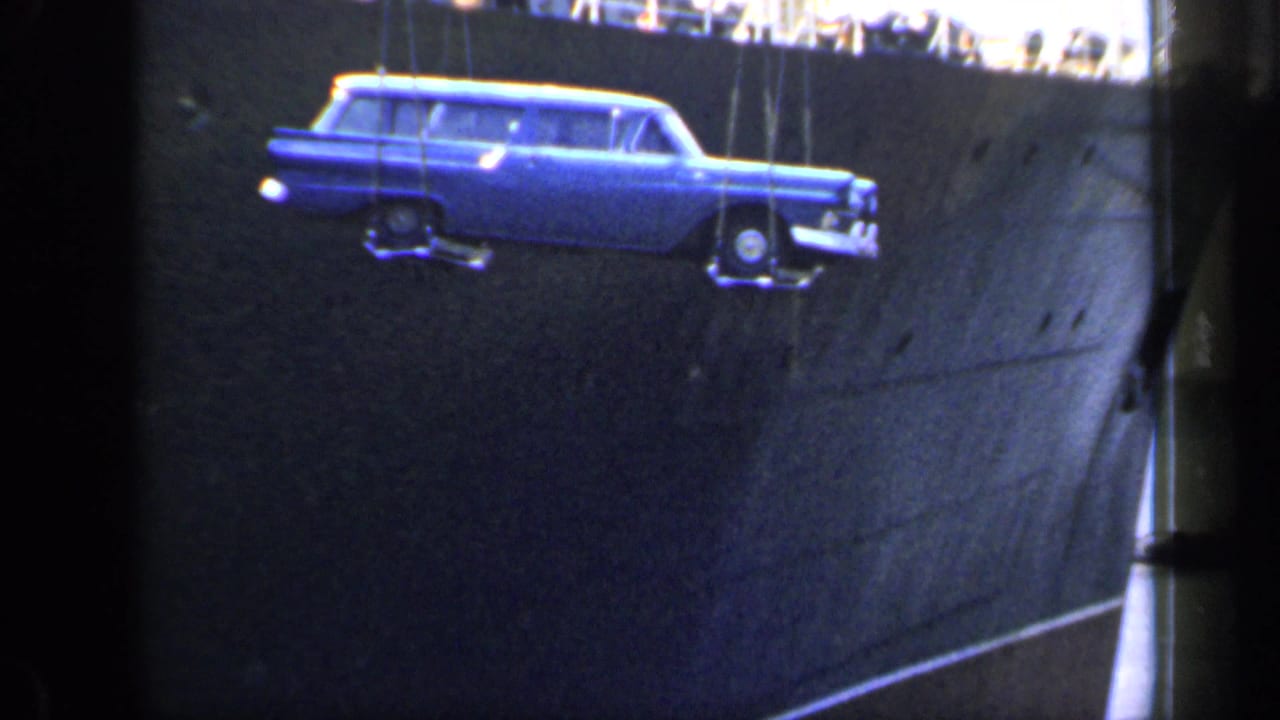 Blue station wagon hangs in front of the dark side of a large ship, held by crane cables.