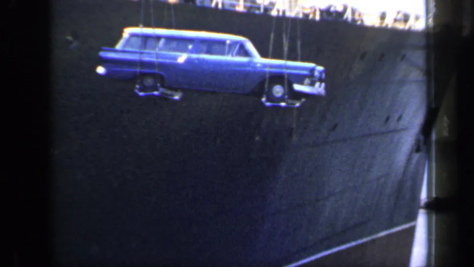 In the 1949 England port footage, the suspended blue station wagon is centered against the dark hull of a large ship.