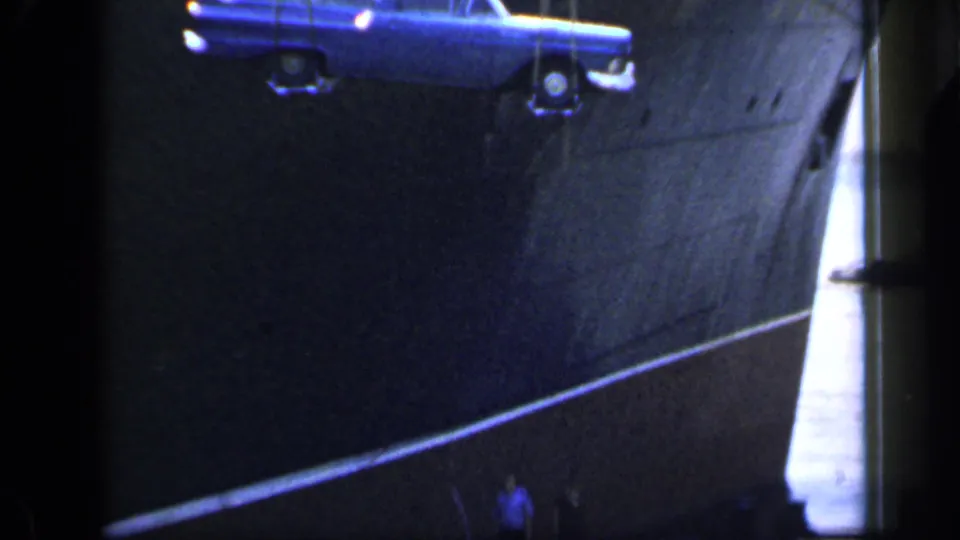 A blue station wagon is lowered beside the dark ship hull, with small dockside people visible below in the 1949 England scene.
