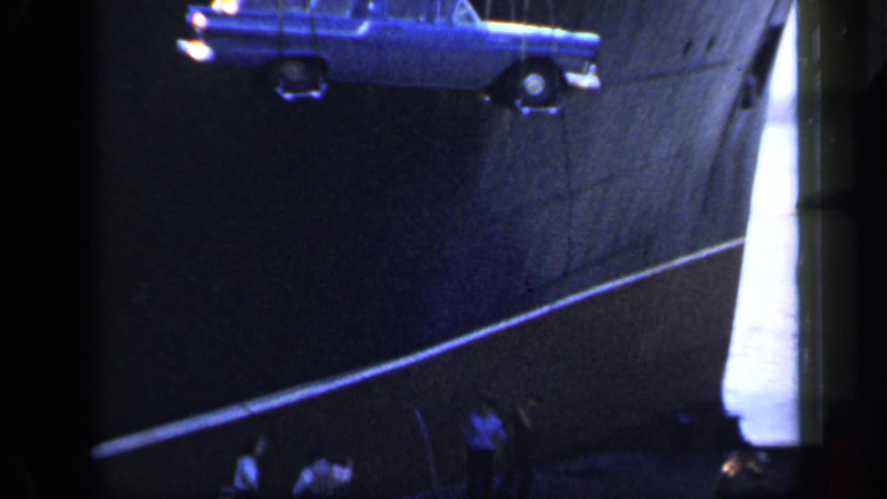 In 1949 England home-movie footage, a blue station wagon hangs from a crane beside a large ship as men watch from the dock.