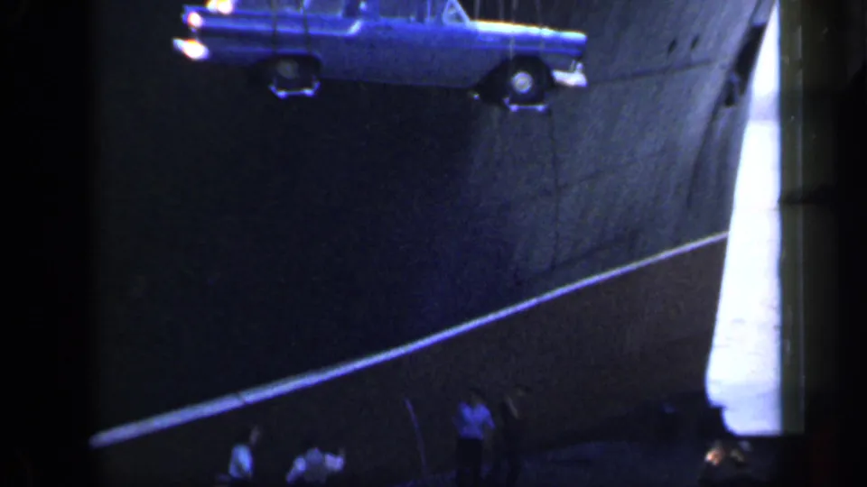 In 1949 England home-movie footage, a blue station wagon hangs from a crane beside a large ship as men watch from the dock.