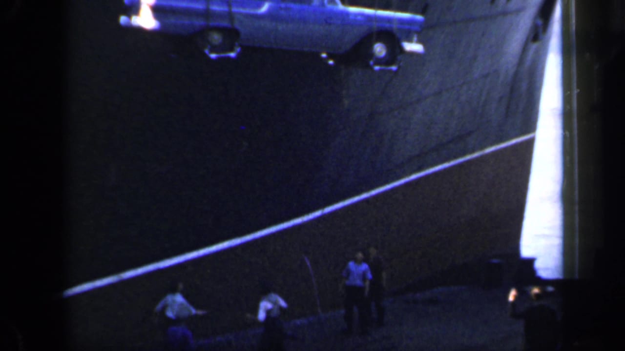 The blue car remains suspended beside the ship, with small figures on the dock emphasizing the scale of the vessel.