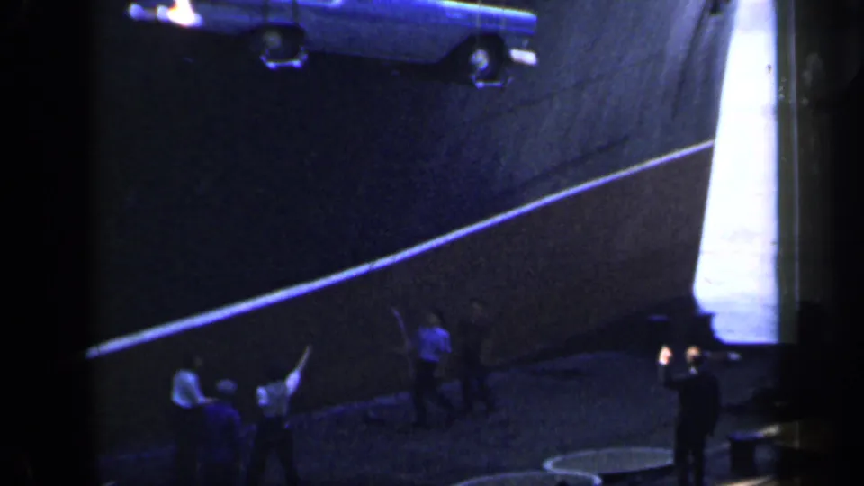 A blue station wagon is lowered from the ship side while men in period clothing gather and gesture on the dock below.