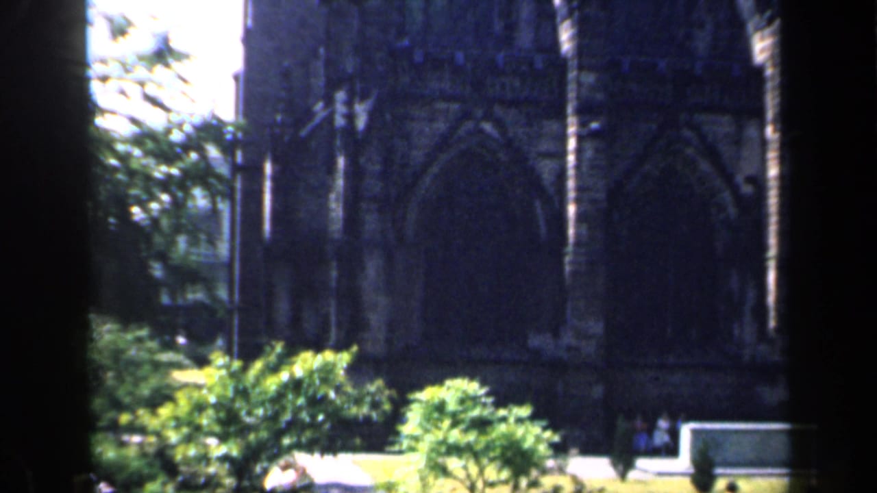 1949 England 8mm frame showing a dark Gothic church exterior, arched window tracery, bright sky, and greenery in the courtyard.