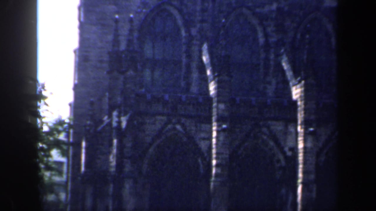 1949 England 8mm frame focused on a Gothic stone facade with tall pointed windows, heavy shadow, and bright overexposed sky at the edge.