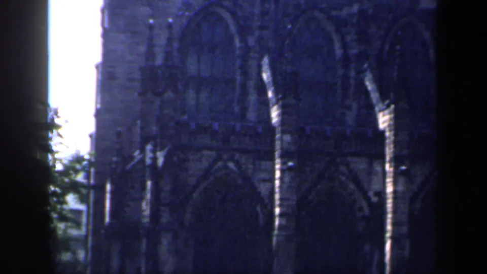 1949 England 8mm frame focused on a Gothic stone facade with tall pointed windows, heavy shadow, and bright overexposed sky at the edge.