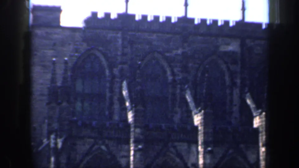 1949 England 8mm frame showing the upper portion of a Gothic church, with crenellated roofline, pointed windows, and blue-shifted stonework.