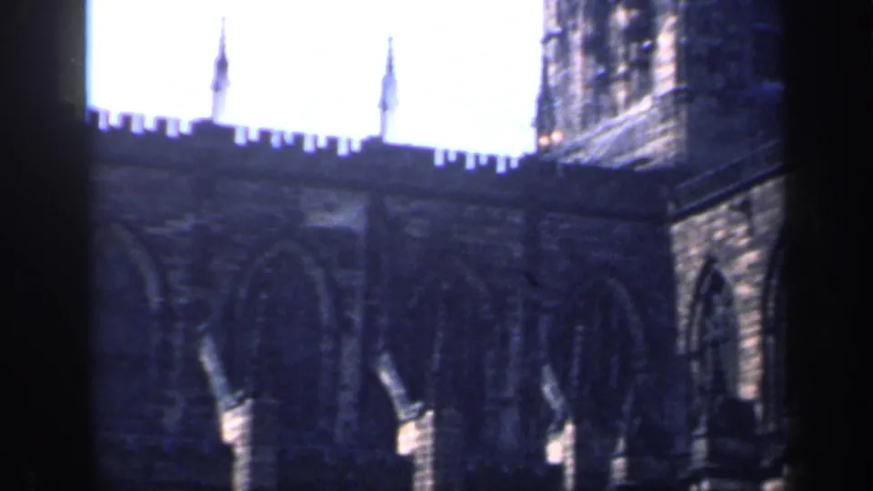 1949 England 8mm frame of a Gothic church exterior, with crenellations, slender roof spires, arched stonework, and a bright washed-out sky.
