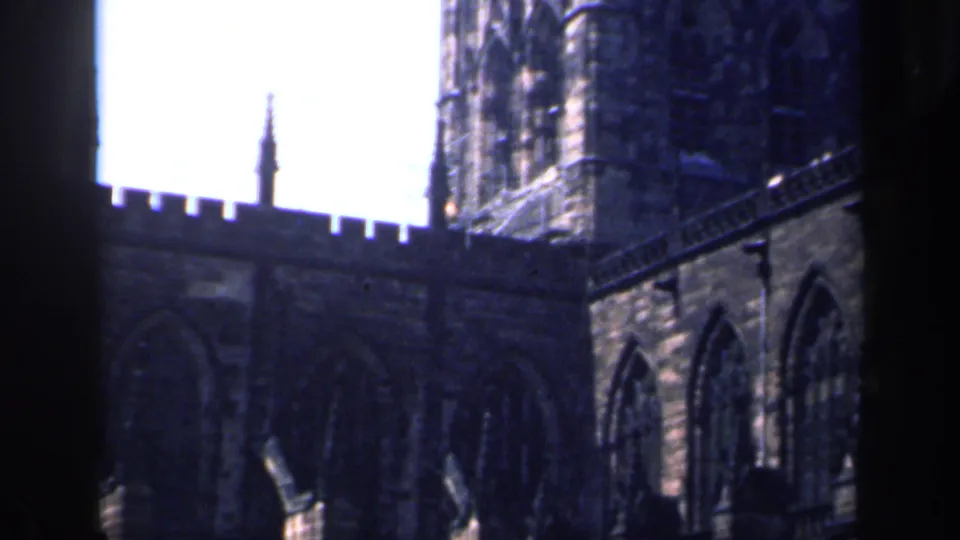 1949 England 8mm frame showing a Gothic stone church tower, pointed windows and crenellated roofline viewed from below.