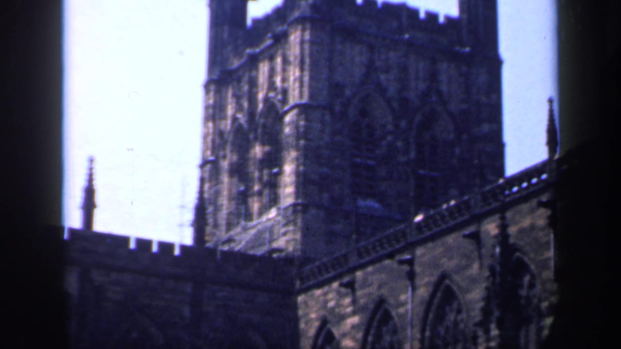 1949 England 8mm frame with a Gothic church tower, arched windows, crenellations and a small spire under bright sky.