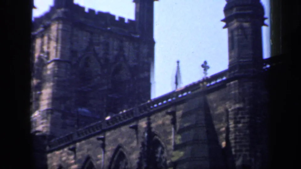 1949 England 8mm frame showing the side of a Gothic stone church with tower, arched openings and decorative roofline.
