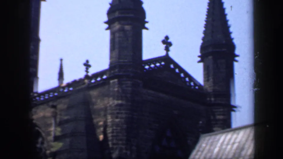 1949 England 8mm frame showing a Gothic church corner with spires, roof finials, stone wall and partial arched window.