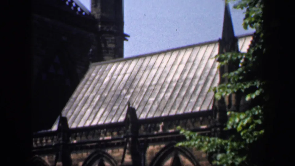 1949 England 8mm view of a church metal roof, pointed spire, upper stone wall, and leaves at the right edge.