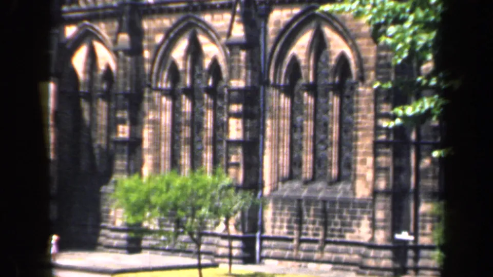 1949 England 8mm view of a sunlit church facade with pointed arched windows, shrubs, trees, and a low fence or wall.