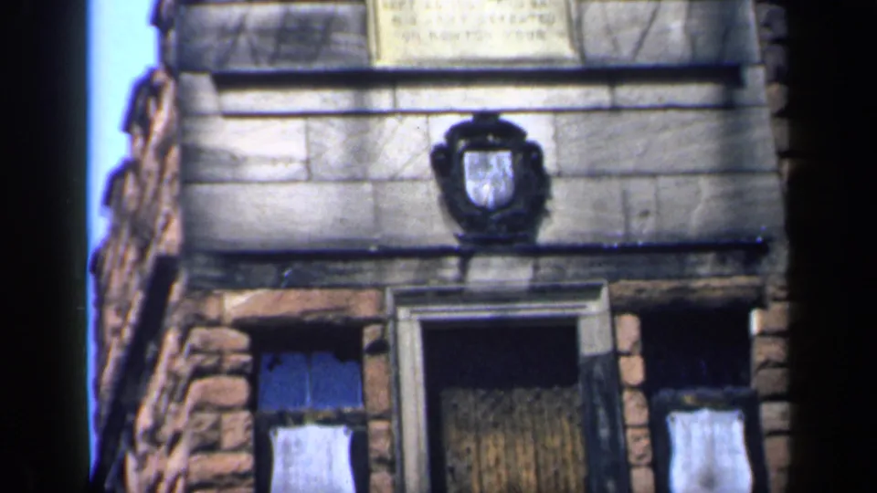 The camera tilts lower across the 1949 England stone building, revealing more of the doorway and boarded windows below the emblem.