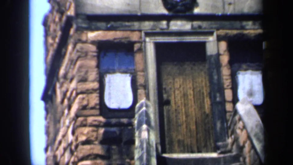 Lower facade detail of the English stone building shows a wooden door, boarded windows, shield emblem, rough masonry, and a stair rail.