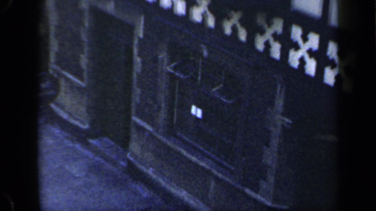 1949 England 8mm frame showing a shadowed doorway, small illuminated window, and patterned timber-framed exterior detail.