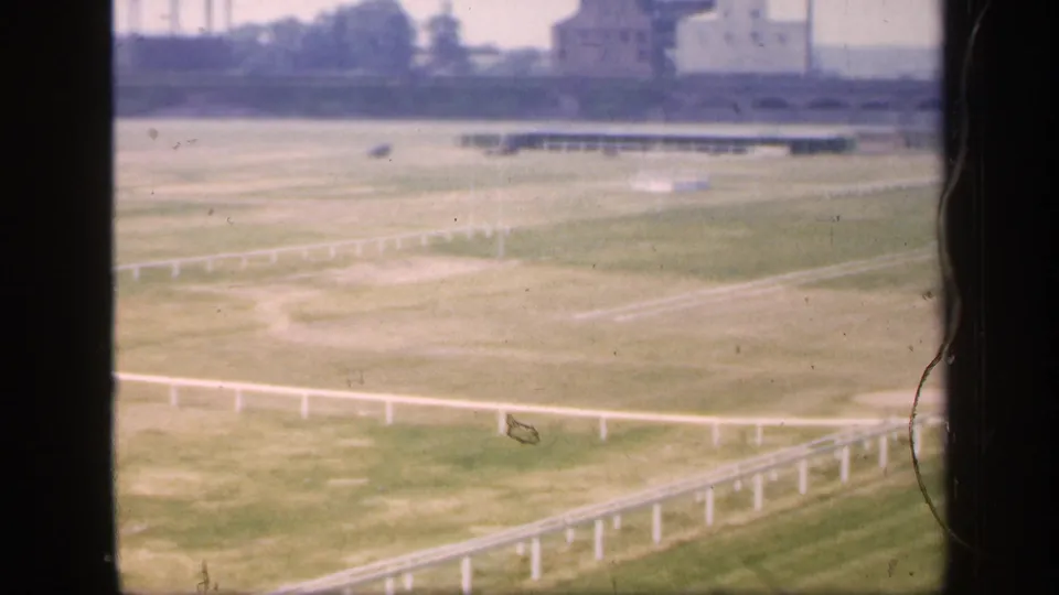 1949 England 8mm frame showing a grassy field with white railings, distant buildings, and trees under a hazy sky.