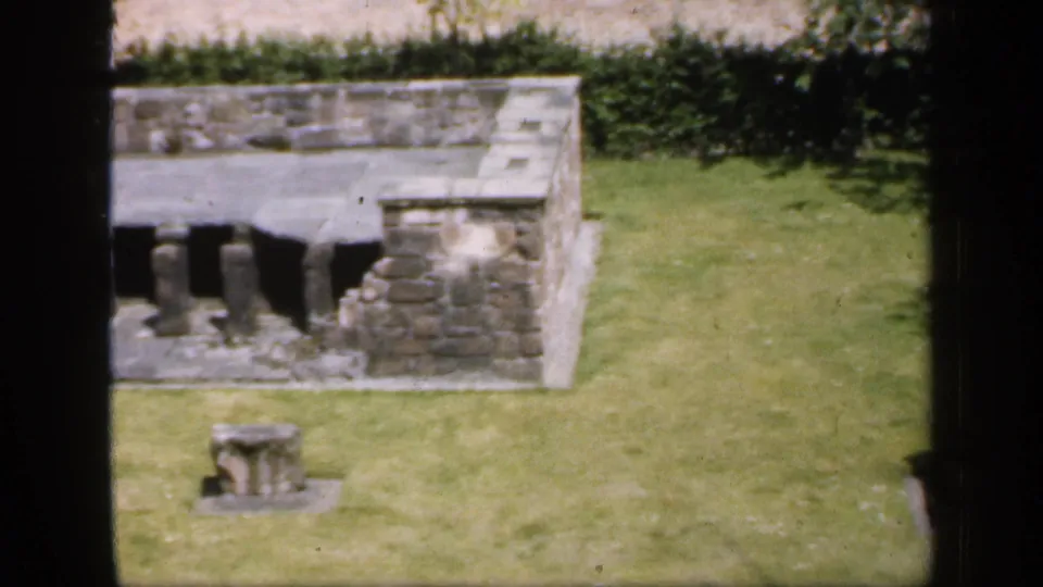 A 1949 England frame looks down past the window to a lawn with a low stone structure and a small column-like remnant.