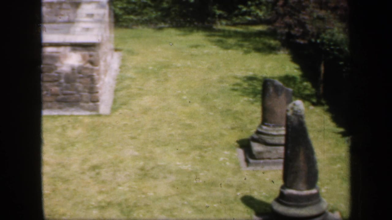 1949 England 8mm frame showing weathered stone ruins, upright monuments, lawn, and dark film gate edges.