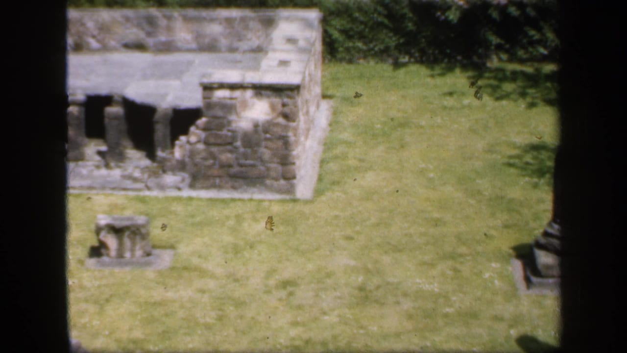 1949 England 8mm frame with a low weathered stone ruin, arched openings, lawn, hedging, and a small stone remnant.