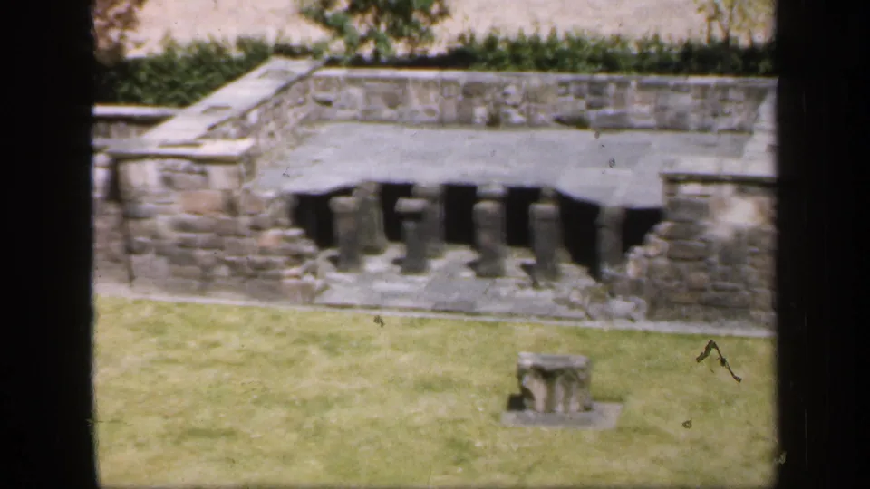 1949 England 8mm frame showing a weathered stone ruin across a lawn, with dark frame edges and a small masonry fragment.