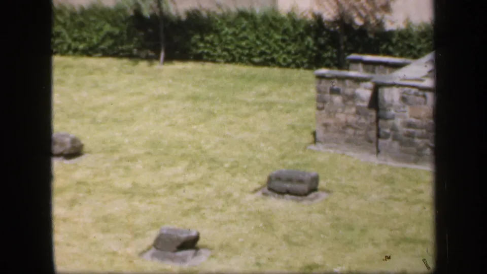 1949 England home-movie frame showing a maintained lawn with scattered stone markers and partial stonework.
