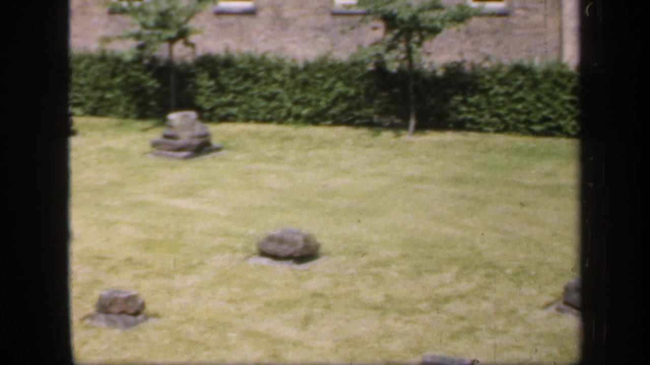 1949 England 8mm frame showing a green lawn with weathered grey stone remnants, clipped hedges, brick wall, and dark window-like borders.