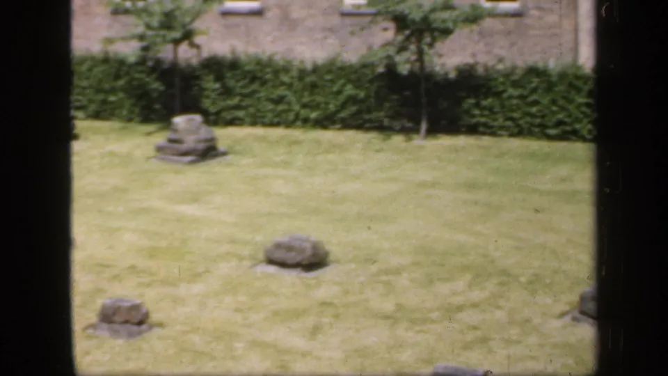1949 England 8mm frame showing a green lawn with weathered grey stone remnants, clipped hedges, brick wall, and dark window-like borders.