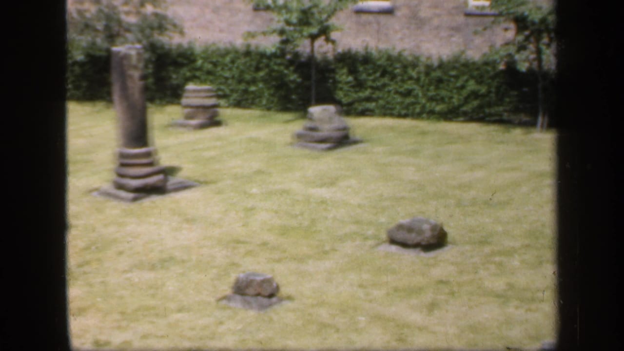 1949 England 8mm frame with a weathered stone column base and smaller grey stone fragments on a sunlit garden lawn.