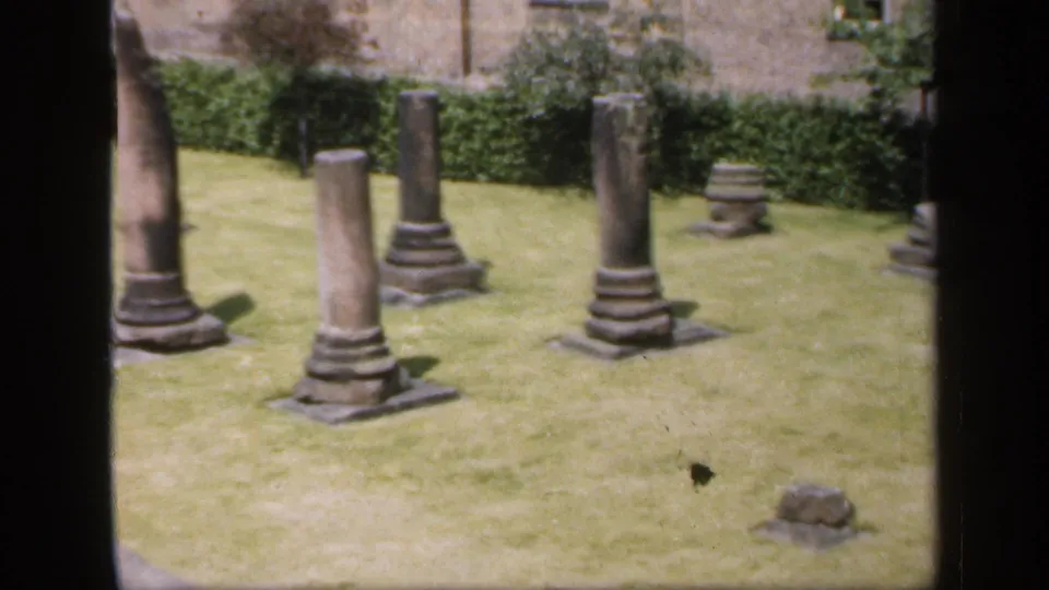 1949 England 8mm frame with three weathered stone columns, low fragments, a green lawn, hedges, and a brick wall in the background.